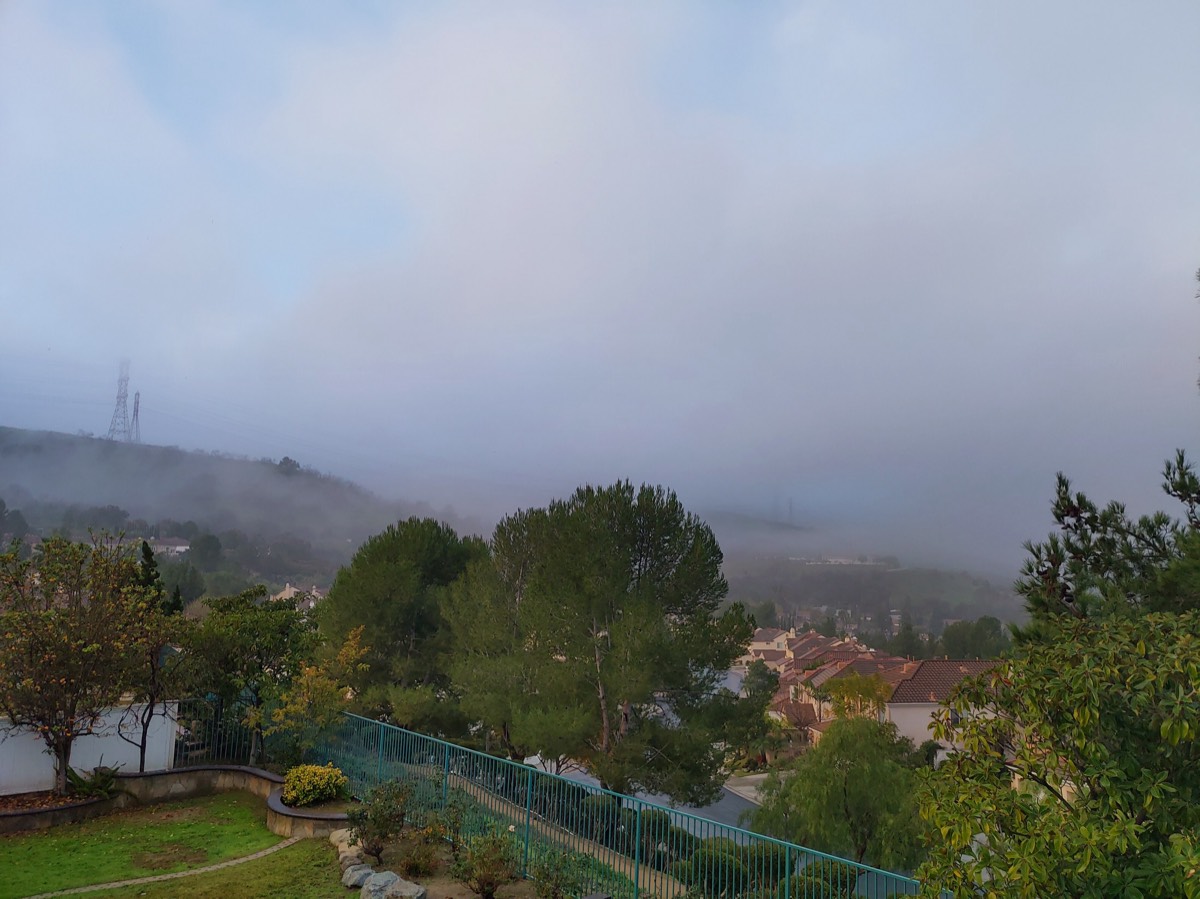 清晨浓雾笼罩的山坡住宅区，高压铁塔与屋顶在雾中若隐若现