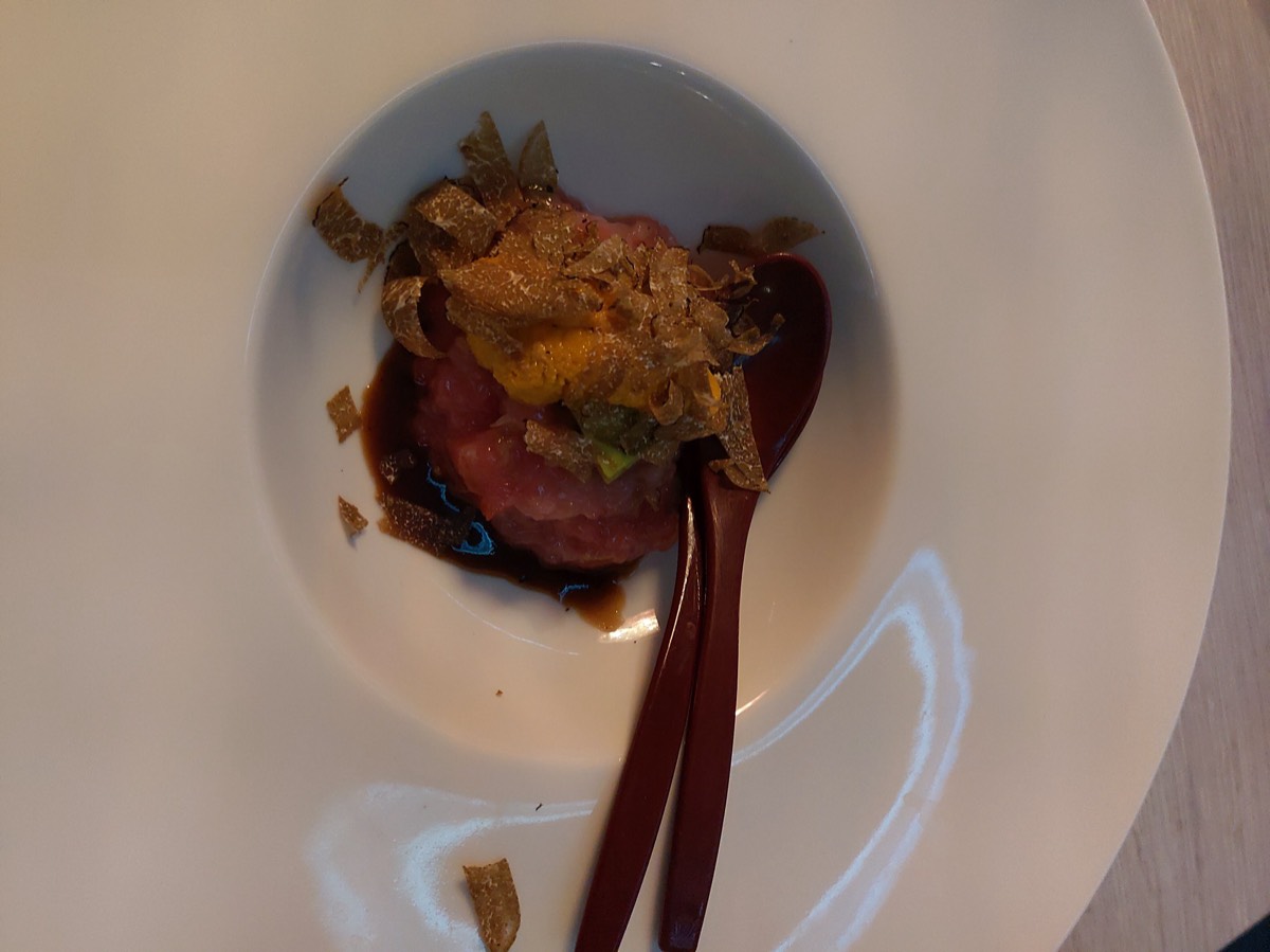白色深盘中的生牛肉塔塔，铺满松露薄片，中央一枚蛋黄，木质勺筷斜放